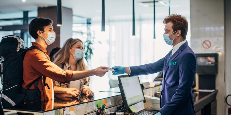 Couple Travelers With Medical Masks On Hotel Reception Talking To Male Receptionist