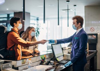 Couple Travelers With Medical Masks On Hotel Reception Talking To Male Receptionist